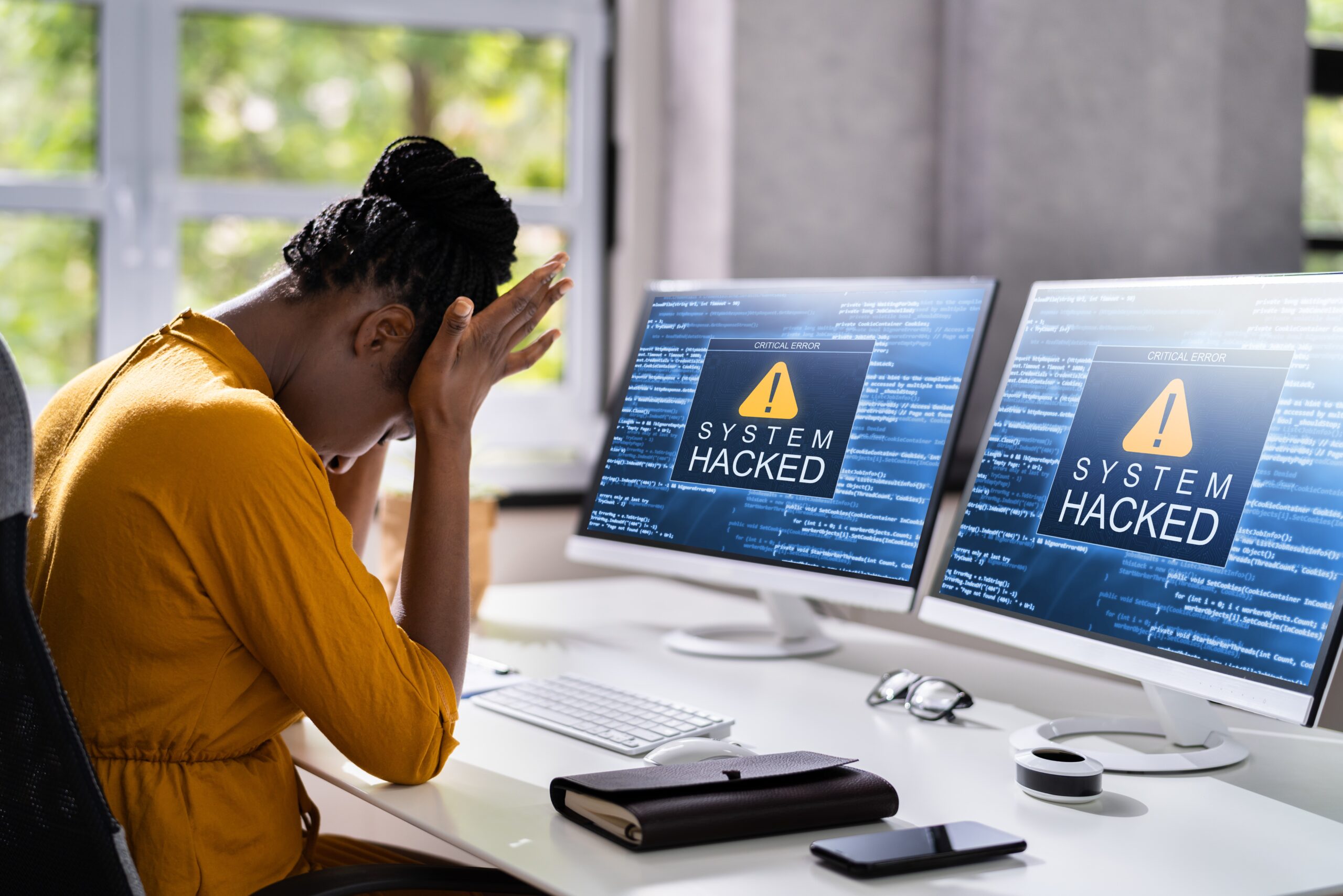 VC cybersecurity concept. Young black woman with head in hands staring at dual monitors saying, 'System hacked'