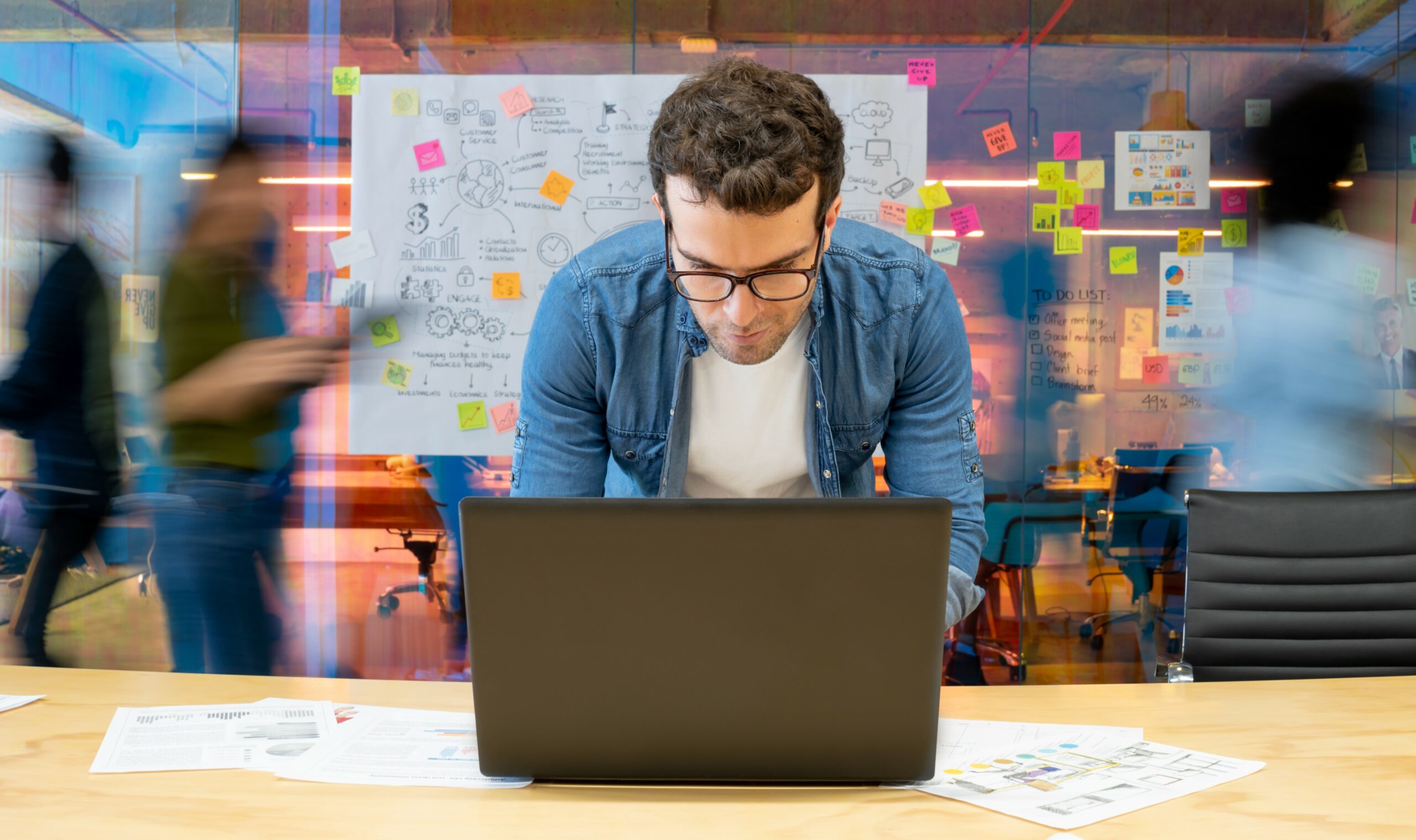 Hiring a contractor concept. Young bespectacled man in denim shirt porting over laptop with agile chart in background
