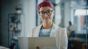 Accounting for entrepreneurs concept. Bespectacled woman with pink hair wearing glasses smiles at camera from behind laptop